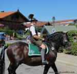 Ein Mann in Lederhose und Gamsbart am Hut reitet auf einem schwarzen Pferd