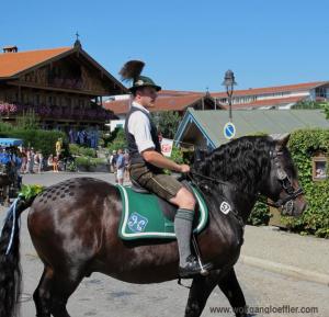 Ein Mann in Lederhose und Gamsbart am Hut reitet auf einem schwarzen Pferd